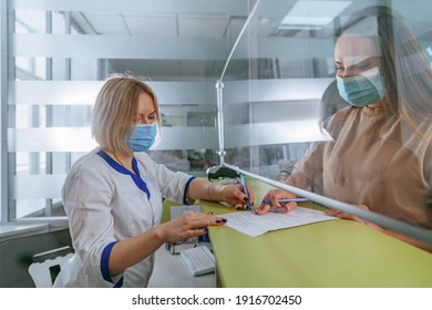 Female nurse explaining patient how to fill form at hospital reception. Doctor and patient wearing protective face masks and using protective glass screen. New normal during Pandemic COVID-19 concept.