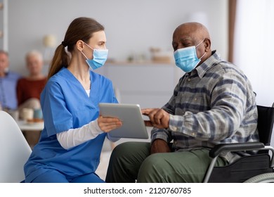 Female Nurse Helping Black Senior Man In Weheelchair Using Digital Tablet, Elderly African American Male Patient Surfing On Internet At Nursing Home, Wearing Protective Face Masks, Copy Space