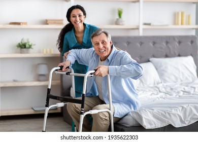 Female nurse helping elderly male with walking frame stand up from bed at home. Professional care for disabled patients