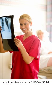 Female Nurse Holding Up An X-ray And Smiling At The Camera With A Senior Female Patient Lying In A Hospital Bed In The Background