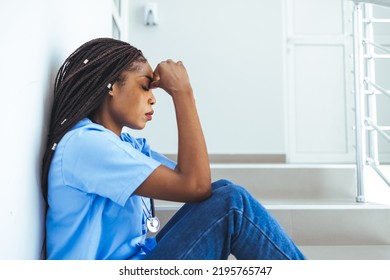 Female Nurse Sitting On The Floor And Looking Distraught. Exhausted Sad Black Doctor Feels Burnout Stress. Stressed Female Medical Taking Break From Work. Being An Intern Is Tough Work