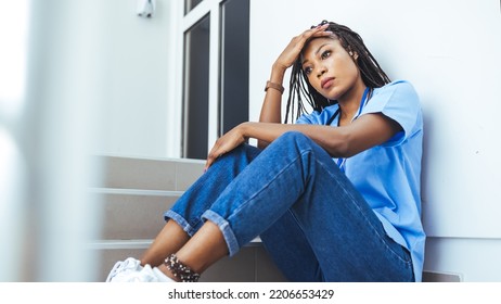 Female Nurse Sitting On The Floor And Looking Distraught. Exhausted Sad Black Doctor Feels Burnout Stress. Stressed Female Medical Taking Break From Work. Being An Intern Is Tough Work