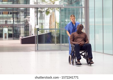 Female Nurse Wearing Scrubs Wheeling Patient In Wheelchair Through Lobby Of Modern Hospital Building - Powered by Shutterstock