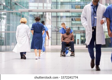 Female Nurse Wearing Scrubs Wheeling Patient In Wheelchair Through Lobby Of Modern Hospital Building