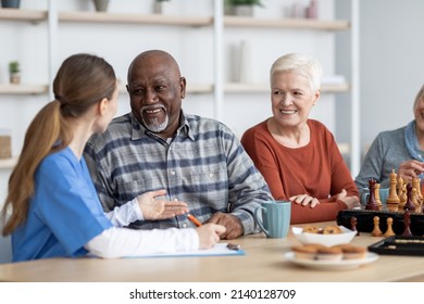 Female Nurse In Workwear Having Conversation With Cheerful African American Senior Man, Multiracial Pensioners Spending Time At Nursing House, Playing Table Games, Drinking Tea, Eating Cookies