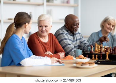 Female Nurse In Workwear Having Conversation With Cheerful Attractive Senior Woman, Multiracial Pensioners Spending Time At Assisted Living, Playing Table Games, Drinking Tea, Eating Cookies