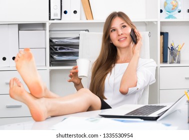 Female Office Worker With Bare Feet On Desk Talking On Phone During Coffee Break 