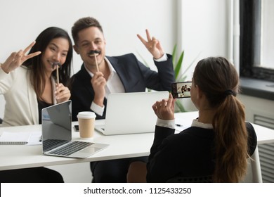 Female office worker taking picture on smartphone of friendly diverse funny smiling colleagues posing together with fake mustache at workplace, coworkers have fun at work break or during staff party - Powered by Shutterstock