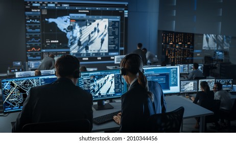 Female Officer Works On A Computer With Surveillance CCTV Video Footage In A Police Monitoring Center With Global Map Tracking On A Big Digital Screen. Employees Sit In Front Of Displays With Big Data