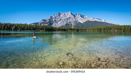 Female On A Stand Up Paddle Board On The Beautiful Clear Two Jack Lake With Mountains Reflecting In The Calm Water On A Summer Day In Banff National Park, Alberta, Canada