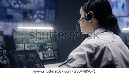 Female operator in headset zooms and monitors security cameras with AI facial recognition, uses tablet. Multiple big screens and PC monitors with CCTV cameras footage. Social safety concept. Close up.