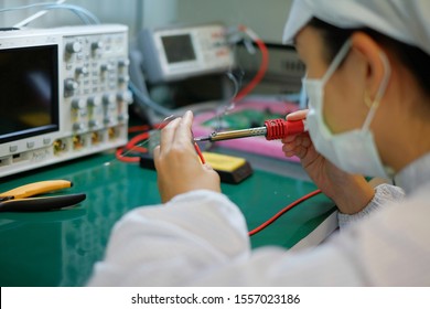 A Female Operator In White Smock (clean Suit), A Cap And A Mask At An Electronic Assembly Line Soldering Copper Wire To A Circuit Board With Solder Iron And Smoke.   