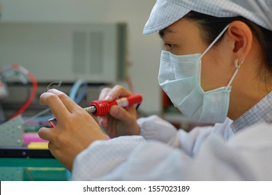 A Female Operator In White Smock (clean Suit), A Cap And A Mask At An Electronic Assembly Line Soldering Copper Wire To A Circuit Board With Solder Iron And Smoke.   