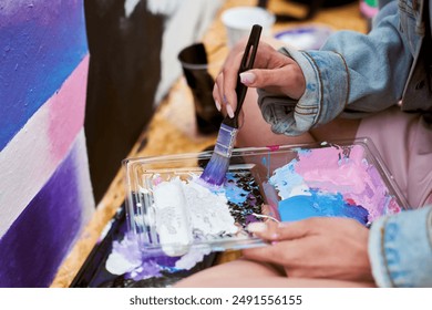 Female painter hand dips paintbrush into palette of colorful paints for live painting of picture for outdoor street exhibition, close up view of female artist hand holding paintbrush - Powered by Shutterstock