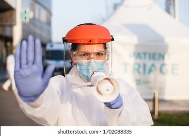 Female Paramedic In Personal Protective Equipment PPE Holding Megaphone Shouting Urgent Commands,standing In Front Of Hospital Outdoor Patient Triage Tent,Coronavirus COVID-19 Pandemic Outbreak Crisis