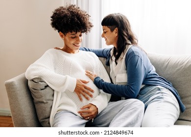 Female parents-to-be sitting together at home. Happy young woman smiling cheerfully while embracing her pregnant wife. Pregnant lesbian couple spending quality time together.