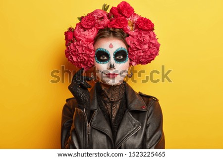 Female participant of Mexican holiday has professional makeup, resembles skull to honor dead relatives has black eyes wears red peonies wreath dressed as spirit models indoor over vivid wall