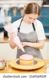 Female pastry chef making delicious cake. Woman putting pink cream from piping bag on sponge layer placed on rotating cake stand in bakery