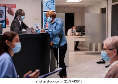 Female Patient With Crutches Talking To Receptionist In Hospital Lobby, Chatting About Medical Appointment And Healthcare. Patient With Physical Impairment At Reception Help Desk.