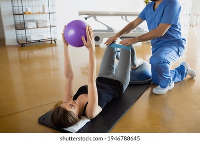 Female patient lying down on a mat, exercising with her physiotherapist using resistance band for legs and a fit ball, in a gymnasium or clinic. Physical therapy concept. - Powered by Shutterstock