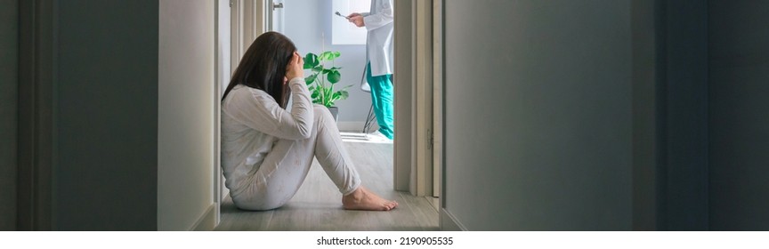 Female Patient In Pajamas Sitting On The Floor In The Corridor Of A Mental Health Center With A Doctor In The Background