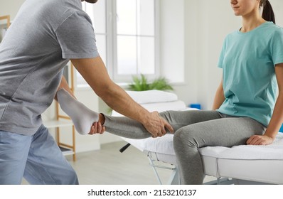 Female Patient Receiving Professional Medical Care In Rehabilitation Center After Leg Injury. Male Physiotherapist Examines Leg And Knee Of Woman Sitting On Medical Couch. Concept Of Medical Care