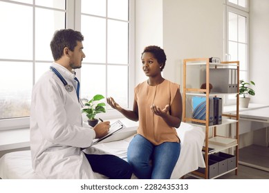 Female patient talking to doctor in his office. Worried young black woman having conversation with male physician in examination room at modern clinic, hospital or medical center - Powered by Shutterstock