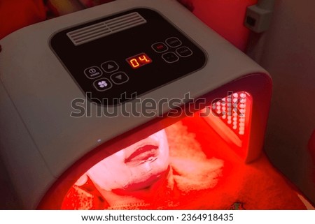A female patient undergoes red LED light or RLT therapy for skin rejuvenation at an aesthetic clinic. Using a phototherapy device with red light wavelength for acne treatments and collagen production.