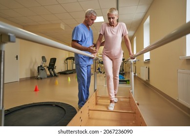 Female Patient Walking Between Parallel Bars At Rehabilitation Room