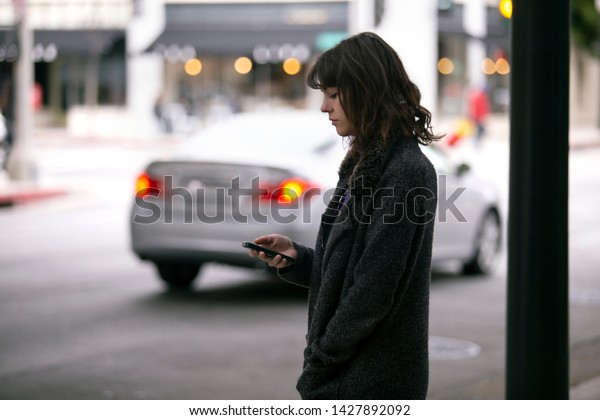 Female pedestrian waiting for a rideshare.  She is\
sharing her gps location via cellphone app so the driver can pick\
her up in the city.  Cars are blurred to obscure make model and\
license plates. 