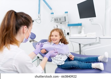 Female pediatric dentist medic making a treatment for adorable little smiling girl sitting in chair in a hospital. Dentist and child in cabinet. Little girl sitting in the dentists office