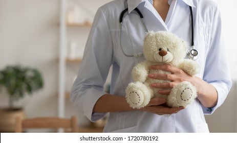 Female pediatrician holding teddy bear in hands. Professional paediatrician doctor wears white coat standing with toy. Children healthcare paediatric medical services background concept. Close up view - Powered by Shutterstock