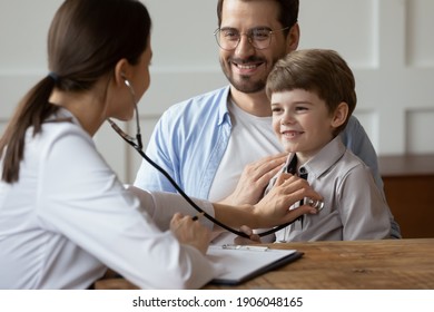 Female Pediatrician Listen To Smiling 6s Boy Patient Heart Chest With Stethoscope During Visit With Dad To Clinic. Woman Doctor Examine Small Child At Consultation With Father. Healthcare Concept.