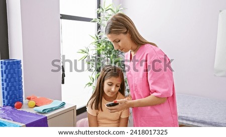 A female pediatrician in a pink scrub shows a device to a young female patient in a bright clinic room.