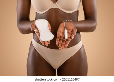 Female Period And Intimate Hygiene Concept. Closeup Cropped View Of Black Woman In Beige Underwear Holding Tampon Vs Clean Menstrual Pad Near Belly, Standing Isolated On Beige Studio Background