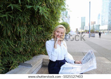 Female person reading newspaper article outside in   with close up face. Concept of mass media and business clothes. Beautiful blonde woman resting near green plant.