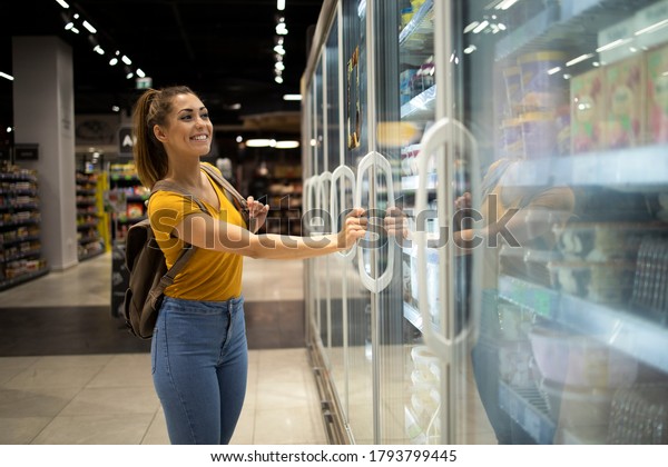 Female\
person with shopping cart opening fridge to take food in grocery\
store. Woman buying groceries in\
supermarket.