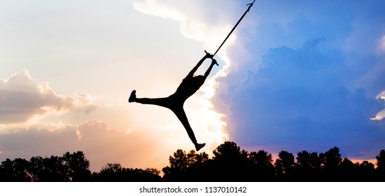 female person silhouette flying in sky, silhouette girl flying on a rope, human  female gymnast hanging on rope against the background of clouds and sunset