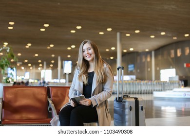 Female person sitting in airport hall with tablet near valise and wearing grey coat. Concept of traveling abroad, modern technology and gladden passengers. - Powered by Shutterstock