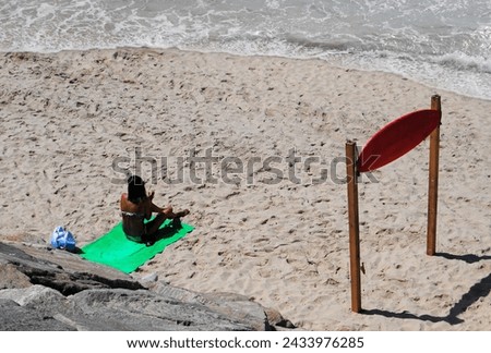 Female person sitting on the beach waiting to see surfers in the sea, green towel, beach entry with surf board