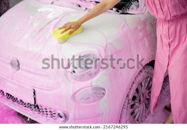 Female person wiping\
car covered in pink nano foam with yellow sponge at car wash.\
Close-up view on car\
hood