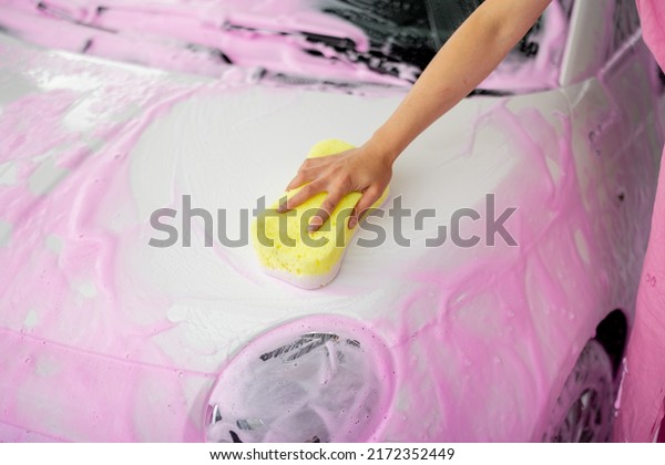 Female person wiping\
car covered in pink nano foam with yellow sponge at car wash.\
Close-up view on car\
hood