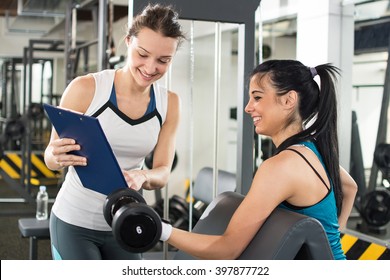 Female personal trainer helping young woman on her work out routines in gym. Fitness woman lifting weights and her personal trainer showing fitness report on a clipboard.