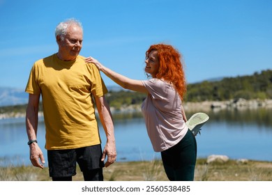 Female personal trainer supports a senior man stretching his leg by a lake, promoting healthy aging and outdoor fitness - Powered by Shutterstock