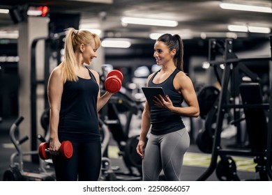 A female personal trainer is tracking progress on tablet while sportswoman is lifting weights in a gym. - Powered by Shutterstock