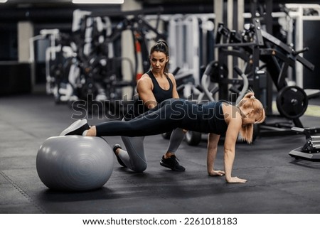 A female personal trainer is training fit sportswoman who is doing exercises with fitness ball in a gym.