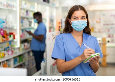 Female Pharmacist In Face Mask Standing In Salesroom Of Drugstore And Holding Notebook In Hands.