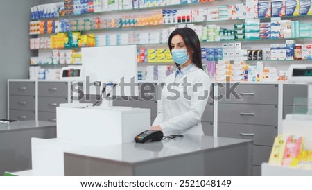 Female pharmacist looking at computer screen. Woman pharmacist working with inventory. Woman in gown and protective mask at background of medicine packaging. Caucasian pharmacist gazing at camera.