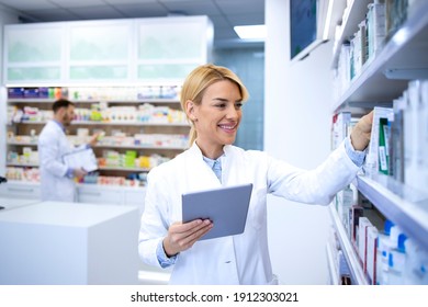 Female Pharmacist Working In Chemist Shop Or Pharmacy On Tablet Computer. Arranging Medicines On The Shelf.