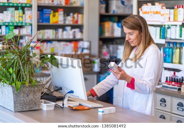 Female pharmacist working in chemist shop or pharmacy.\
Pharmacist using the computer at the pharmacy. Portrait of young\
female pharmacist holding medication while using computer at\
pharmacy counter 